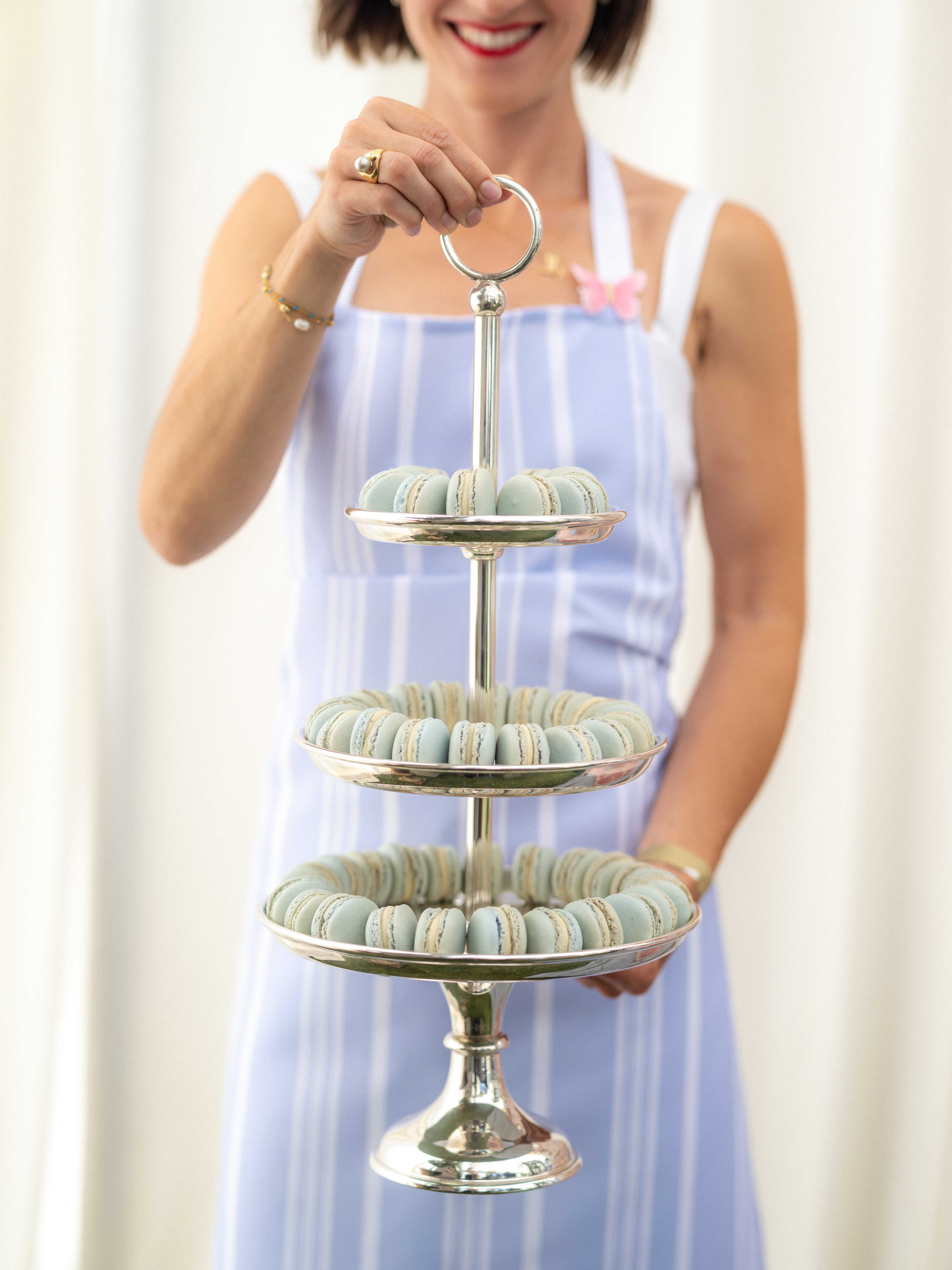 Woman holding a silver three-tiered stand with pastel macarons against a white background. Large, customized macaron dessert bar set up at a premiere Santa Barbara wedding venue, providing a memorable guest experience. Photo by Elizabeth Messina