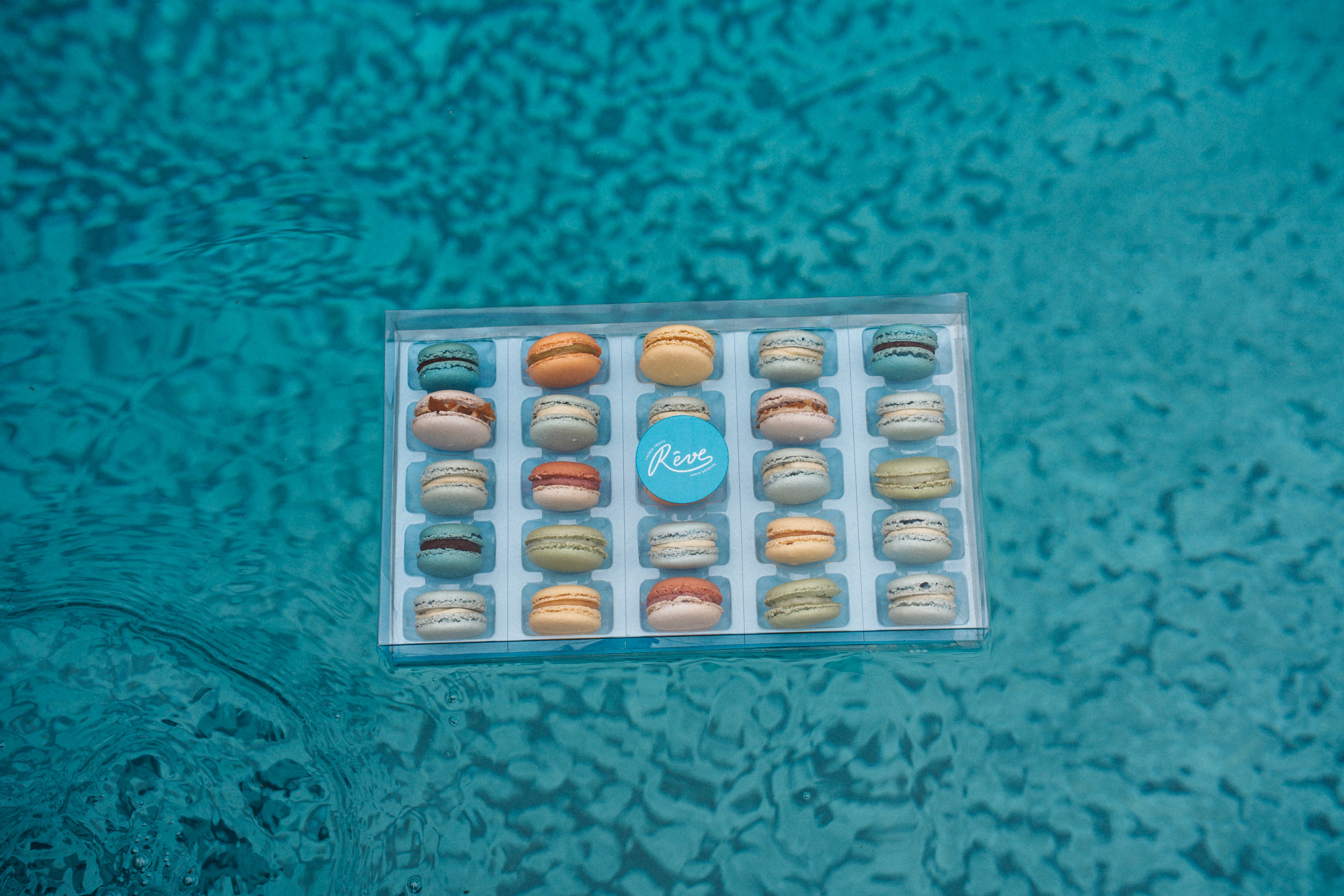 Box of 25 French macarons with a blue label on a textured teal surface