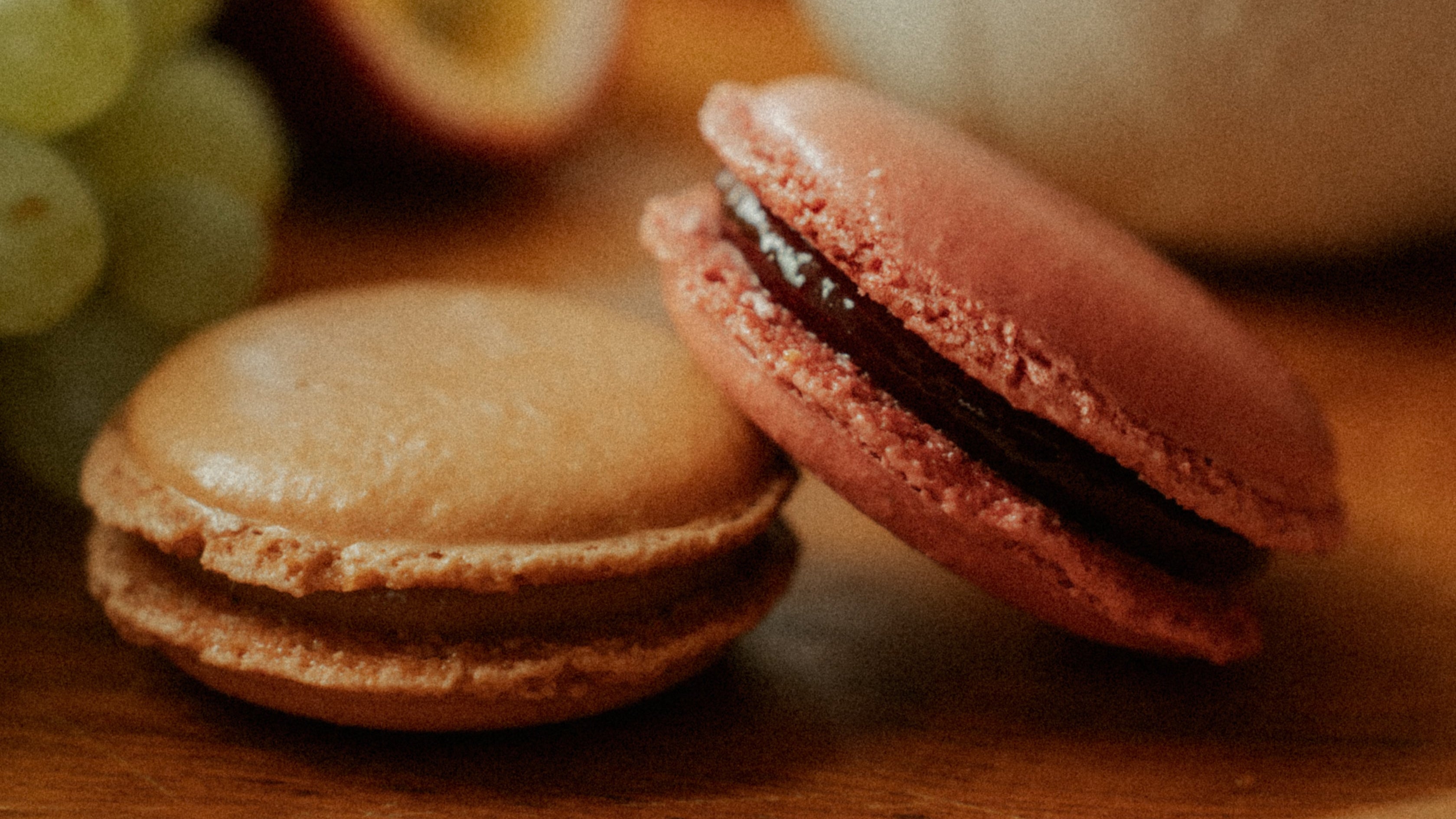 Two macarons on a wooden plate with grapes and an orange in the background.