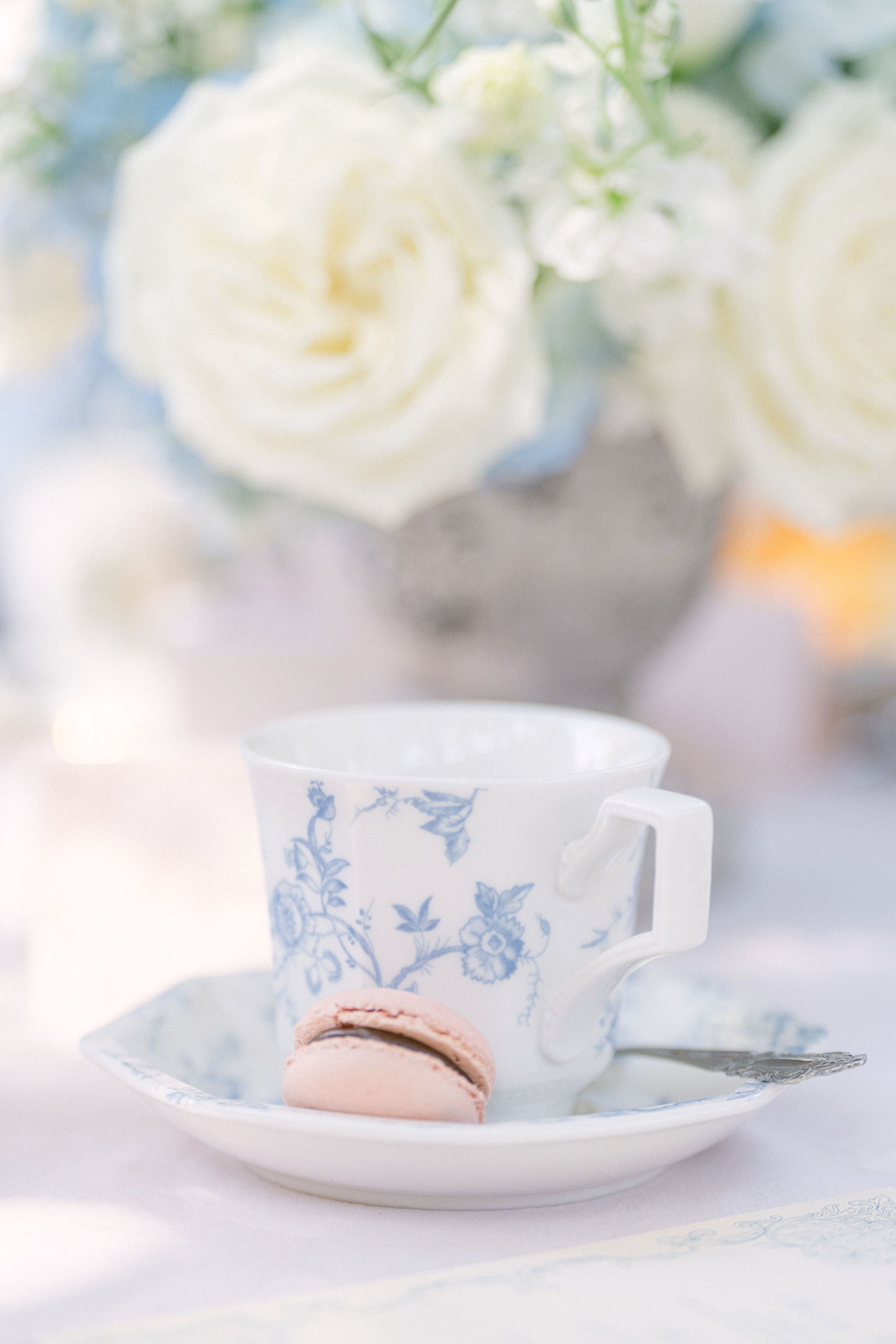 Teacup with floral design on a saucer with a pink macaron, blurred flowers in the background