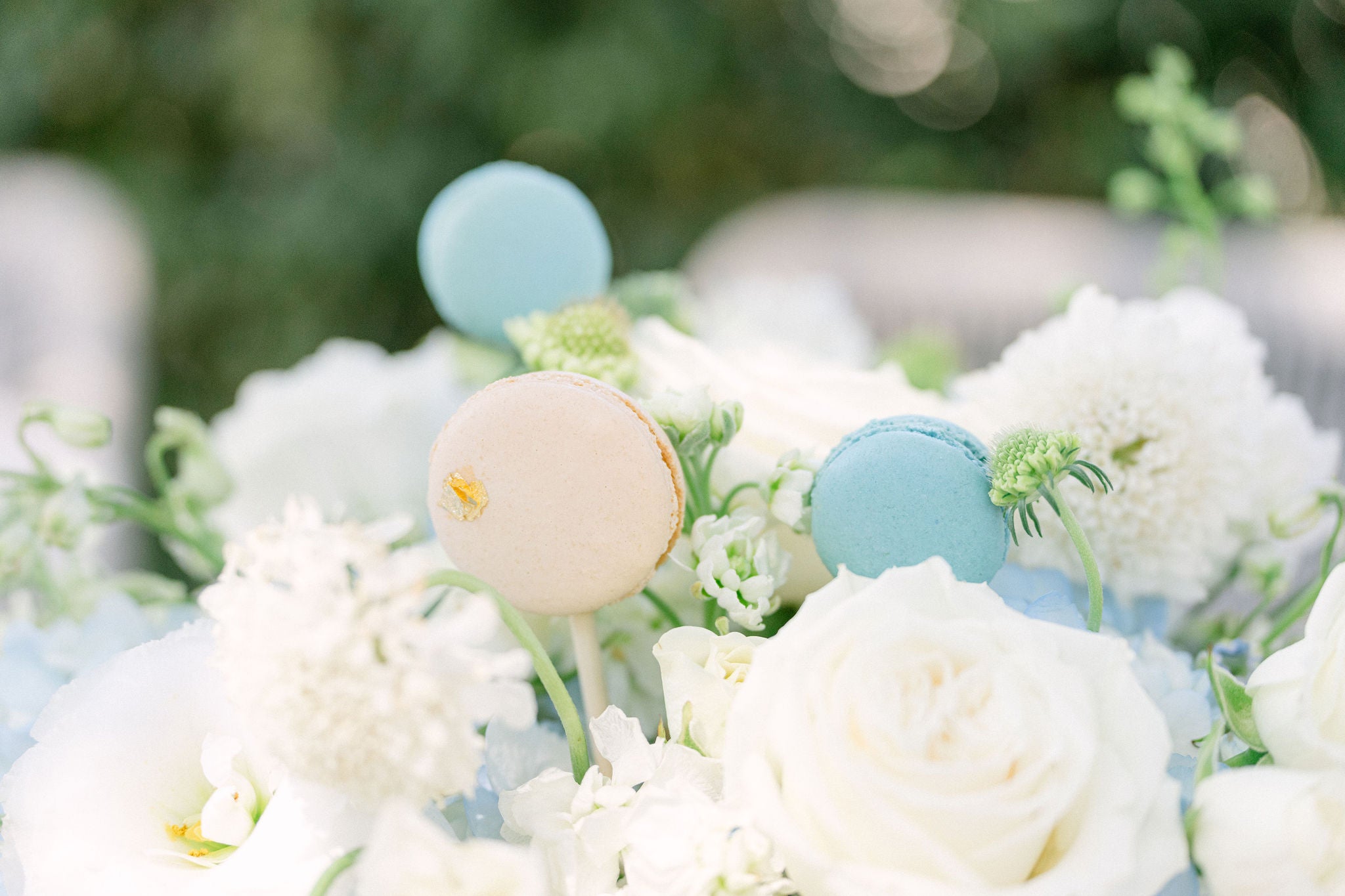 Decorative arrangement with pastel-colored macarons and white flowers on a blurred green background. Bespoke macaron display with custom color palette and 24K gold leaf, designed for a luxury Montecito wedding.