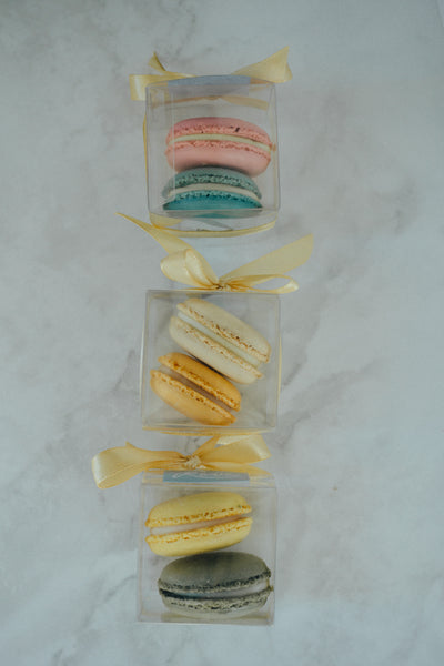 Assorted colorful macarons in a clear box with a yellow ribbon on a marble surface.