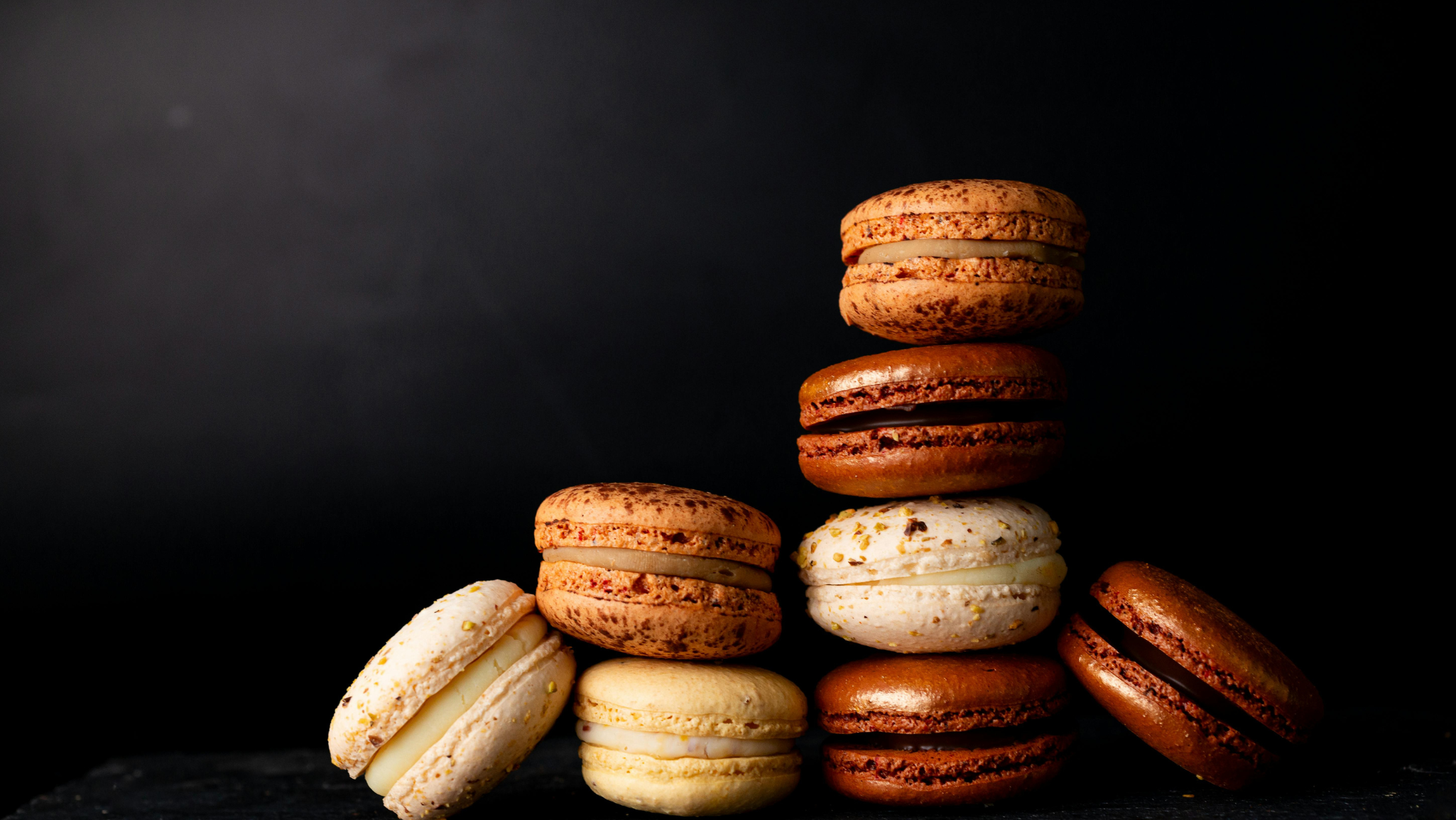 A group of assorted French macarons in various colors, including brown, beige, and yellow, stacked and arranged on a dark black background.