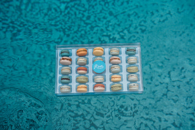 Box of 25 French macarons with a blue label on a textured teal surface
