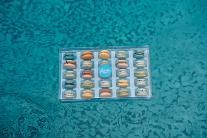 Box of 25 French macarons with a blue label on a textured teal surface