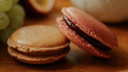 Two macarons on a wooden plate with grapes and an orange in the background.