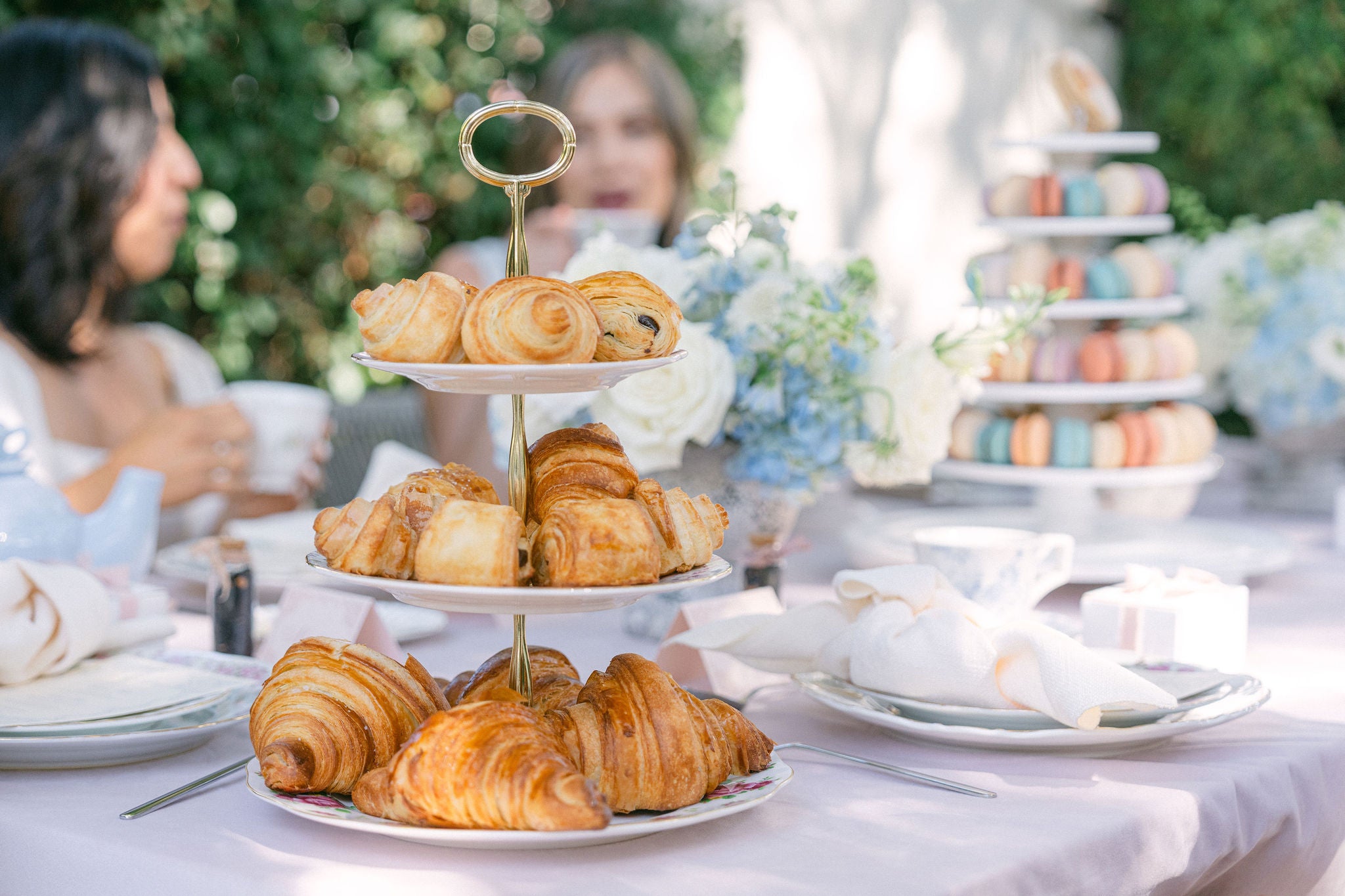 Three-tiered stand with pastries on a table outdoors with people in the background. Artisanal French Croissants | Local Santa Barbara Delivery - Garden parties and baby showers