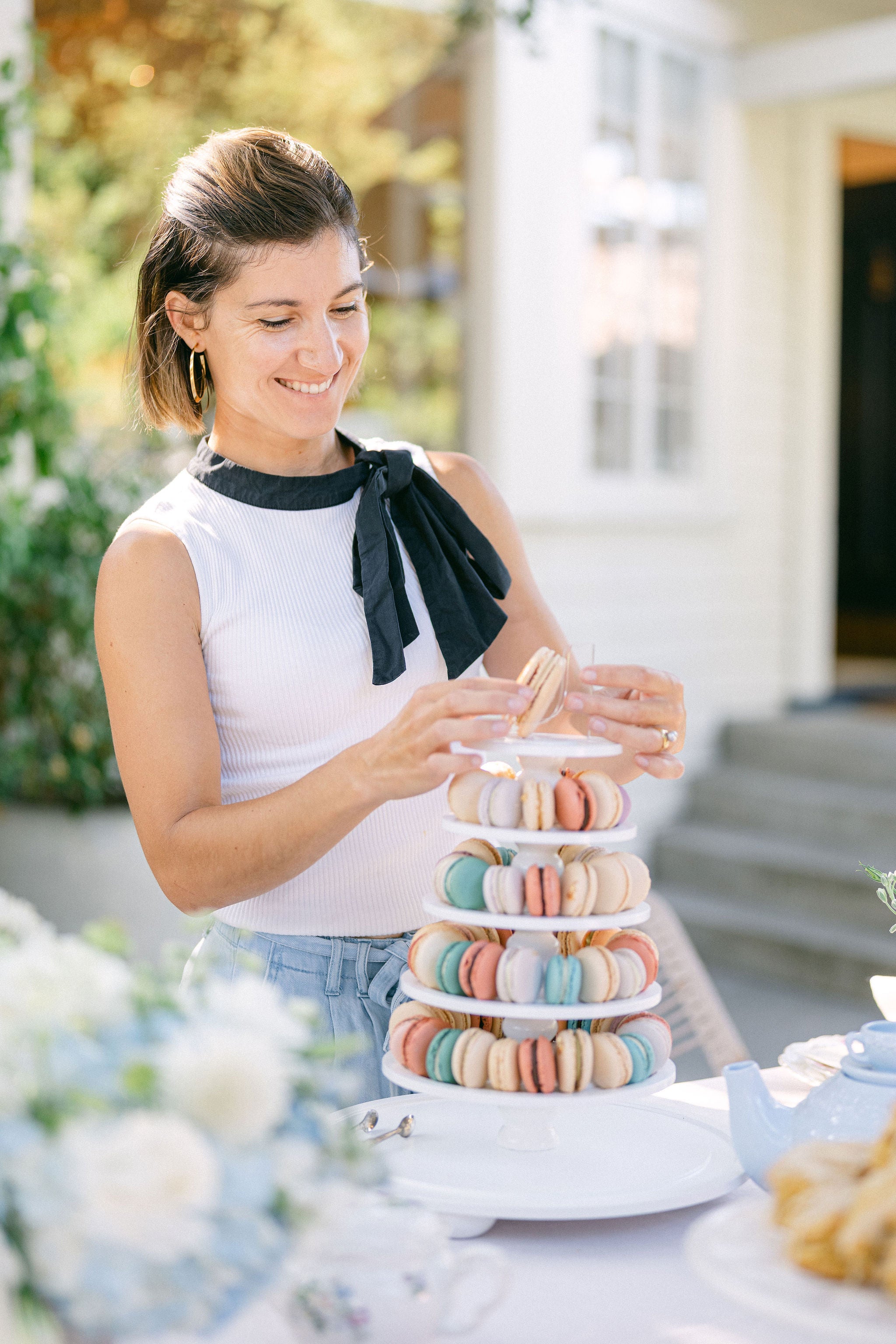 Woman arranging macaron cookies on a tiered stand outdoors.